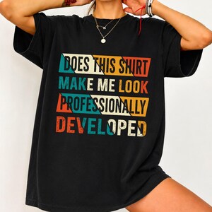 May include: A black short-sleeve t-shirt featuring a graphic with the text DOES THIS SHIRT MAKE ME LOOK PROFESSIONALLY DEVELOPED. The text is arranged in four horizontal rows with a retro-inspired colour palette of orange, teal, and cream.