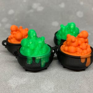 May include: Four miniature black cauldrons with orange and green bubbling liquid inside. The cauldrons are made of plastic and have a Halloween theme.