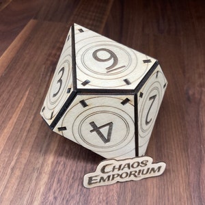 May include: A wooden, multi-sided dice with the numbers 3, 4, 7, and 9 printed on the visible faces. The dice is sitting on a wooden surface with a small sign that reads "Chaos Emporium".