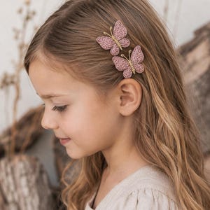 May include: Two small butterfly hair clips featuring dusty pink crocheted wings and a metallic gold wire body. The butterfly wings have a textured, woven appearance, and the gold wire forms the central body and antennae of each insect-shaped accessory.