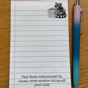 May include: A white lined notepad featuring a black and white illustration of a raccoon wearing sunglasses and holding a stack of pizza boxes at the top, with the text "Feel those endorphins? Go ahead, cross another thing off your list!" and a blue ombre pen.