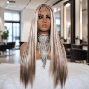 May include: A long, straight wig featuring a blend of platinum blonde and dark brown highlights. The hair is parted in the center and styled with a sleek, smooth texture. A multi-layered, silver-toned fringe necklace hangs around the neck.
