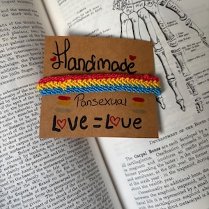 Peut inclure: Un bracelet d'amitié tissé avec des rayures horizontales de fils rose, jaune et bleu. Le bracelet est fixé sur une carte brune portant le texte manuscrit "Handmade", "Pansexual" et "Love = Love" avec de petites illustrations de cœurs rouges.