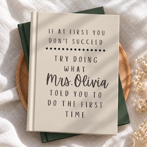 May include: A beige hardcover notebook with black text that reads: IF AT FIRST YOU DON'T SUCCEED, TRY DOING WHAT Mrs. Olivia TOLD YOU TO DO THE FIRST TIME. The notebook is layered on top of a dark green notebook and a round wooden tray.