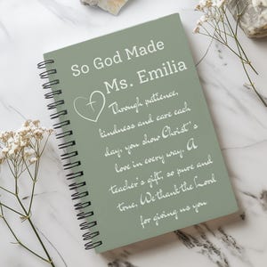 May include: A spiral-bound notebook with a sage green cover. The cover features white text that reads "So God Made Ms. Emilia" and a heart symbol. Additional text expresses gratitude for a teacher. The notebook is on a marble surface.