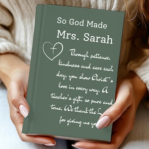 Puede incluir: Un libro verde con texto blanco, sostenido por una persona. El texto dice "So God Made Mrs. Sarah" seguido de un poema sobre el regalo de un maestro. También se ve un corazón con una cruz.