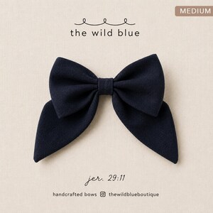 May include: A dark navy blue fabric bow with a textured surface. The text "MEDIUM" appears in a brown box at the top right. Below the bow, the text reads "jer. 29:11", "handcrafted bows", and "thewildblueboutique" with an Instagram icon.