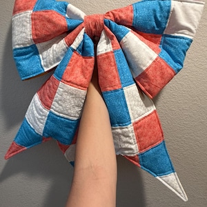 May include: A large, oversized bow constructed from quilted fabric squares in a repeating pattern of coral pink, bright blue, and white. The fabric features subtle floral and geometric prints, with visible stitching lines creating a grid texture.