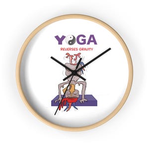 May include: A round wall clock with a light wood frame and a white face. The clock features the purple text YOGA with a yin-yang symbol as the O, and the red text REVERSES GRAVITY. A cartoon illustration shows a creature doing a yoga pose.