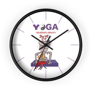 May include: A round wall clock with a black frame and white face. The center features a cartoon illustration of a figure in a headstand on a purple mat. Above the figure, the text reads YOGA REVERSES GRAVITY with a yin-yang symbol as the O.