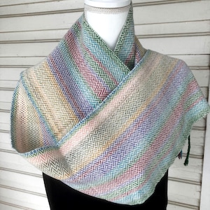 May include: A woven infinity scarf featuring a multi-colored chevron pattern in shades of green, blue, yellow, purple, and pink. The fabric displays a consistent zigzag texture throughout, creating a soft, draped appearance around the neck.