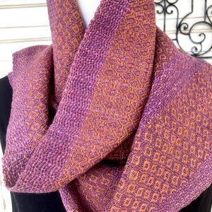 May include: A woven infinity scarf featuring a textured diamond pattern in shades of purple and orange. The fabric displays a dense, interlaced weave with a consistent geometric motif throughout, creating a soft, layered appearance around the neck.