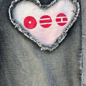 May include: A pair of dark grey denim jeans featuring a large, heart-shaped patch sewn onto the fabric. The patch is light-colored with frayed edges and displays three distinct red circular symbols, each containing white geometric shapes inside.