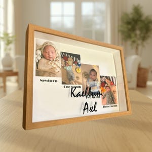 May include: A rectangular light wood shadow box frame containing four photographs of an infant labeled Newborn, One Month, Two Month, and Three Month. The names Kaellum and Axel are displayed in black script lettering on the white background.