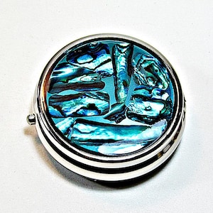 May include: A round, silver-toned metal pill case featuring an inlay of iridescent abalone shell fragments on the lid. The shell pieces display a vibrant mix of blue, green, and teal hues with a polished, reflective finish, set within a smooth frame.