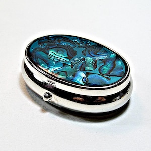 Pode incluir: Uma caixa de comprimidos oval de metal com acabamento prateado polido. A tampa apresenta um mosaico incrustado de peças de concha de abalone iridescente em tons vibrantes de azul profundo, verde-azulado e verde, criando uma superfície cintilante.