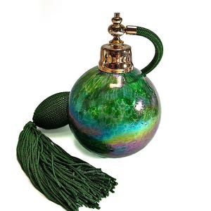 May include: A vintage perfume bottle with a round, iridescent green glass body. It features a gold-colored metal top and a dark green sprayer with a matching tassel. The bottle is designed for dispensing perfume.