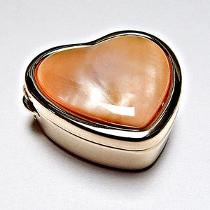 Peut inclure: Une boîte à bijoux en forme de cœur avec une base en métal doré poli et une incrustation lisse et irisée de couleur pêche sur le couvercle. Ce petit récipient présente une conception à charnière avec une finition réfléchissante et brillante.