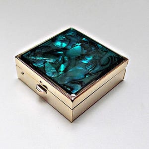 Peut inclure: Une boîte à pilules carrée en métal doré avec un couvercle à charnière incrusté de fragments de coquille d'ormeau bleu-vert irisés. La boîte présente une finition lisse et polie sur les côtés et un petit loquet arrondi sur le bord avant.
