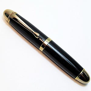 May include: A black fountain pen with a glossy finish, featuring gold-colored metal accents on the cap, the clip, the center band, and the end of the barrel. The pen has a rounded, tapered shape with a smooth, reflective surface throughout.