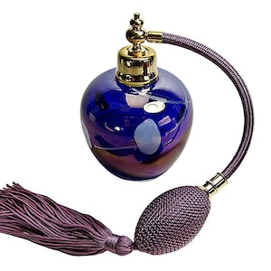 May include: A round, deep blue glass perfume bottle with a gold-toned metal atomiser top. A purple fabric-covered hose connects the bottle to a matching purple textured bulb pump, which features a long, dangling purple tassel at the base.