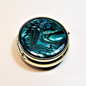 Peut inclure: Un miroir de poche rond en métal doré avec une incrustation de nacre d'ormeau irisée sur le couvercle. La nacre présente des nuances profondes de bleu, de vert et de sarcelle avec une finition polie et brillante. Le boîtier métallique est lisse.