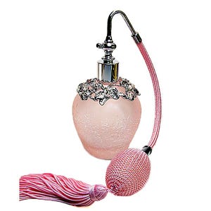 May include: A frosted light pink glass perfume bottle with a textured surface, featuring a silver-toned metal collar with floral detailing, a curved silver nozzle, and a pink mesh-covered bulb pump attached to a long pink decorative tassel.
