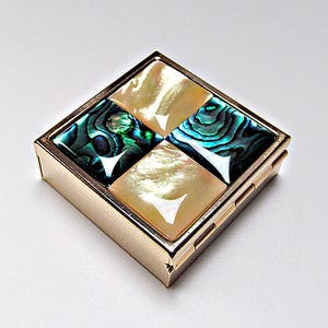 May include: A square gold-toned metal compact case featuring a checkerboard pattern on the lid. The design consists of four alternating squares: two squares of iridescent, dark green and blue abalone shell and two squares of smooth, cream-colored mother-of-pearl.