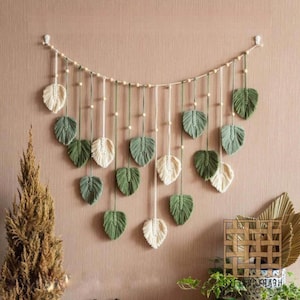 Pode incluir: Uma tapeçaria de macramé com um cordão horizontal decorado com pequenas contas de madeira e múltiplos ornamentos de fibra em forma de folha pendurados em tons alternados de verde sálvia e creme, suspensa por dois ganchos de parede brancos.