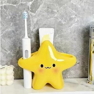 May include: A bright yellow, star-shaped ceramic toothbrush and toothpaste holder featuring a simple, printed face with black eyes, a small curved mouth, and soft pink cheeks. The holder has two circular openings for holding items.