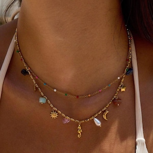 Op de afbeelding: Een goudkleurige gelaagde ketting bestaande uit twee fijne kettinkjes. De bovenste ketting is versierd met kleine, veelkleurige kraaltjes. De onderste ketting bevat diverse bedels, waaronder een zon, een maansikkel, een zeepaardje en een parel.