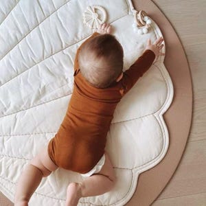 May include: A baby wearing a long-sleeved rust-colored bodysuit lies on a cream-colored, shell-shaped quilted play mat. Two small, circular, light-colored wooden and silicone teething toys are placed near the baby's outstretched hands.