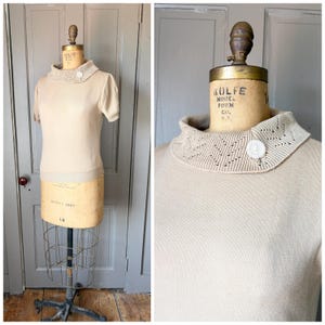 May include: A beige short-sleeved sweater with a decorative, open-knit collar. The collar is fastened with a single white button. The sweater is displayed on a vintage dress form with a metal base.