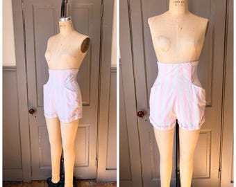 May include: High-waisted shorts featuring a vertical striped pattern in light pink and white. The shorts have a structured waistband, side pockets, and cuffed hems, displayed on a beige dress form mannequin in both side and front views.