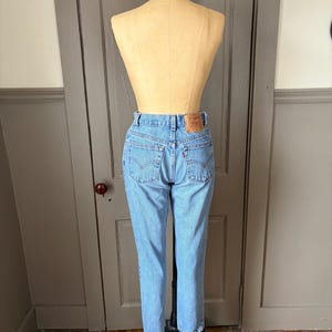 May include: Light blue denim jeans on a mannequin. The jeans have a classic five-pocket design, a button and zipper closure, and a leather patch on the back. The jeans are straight-legged and appear to be vintage.