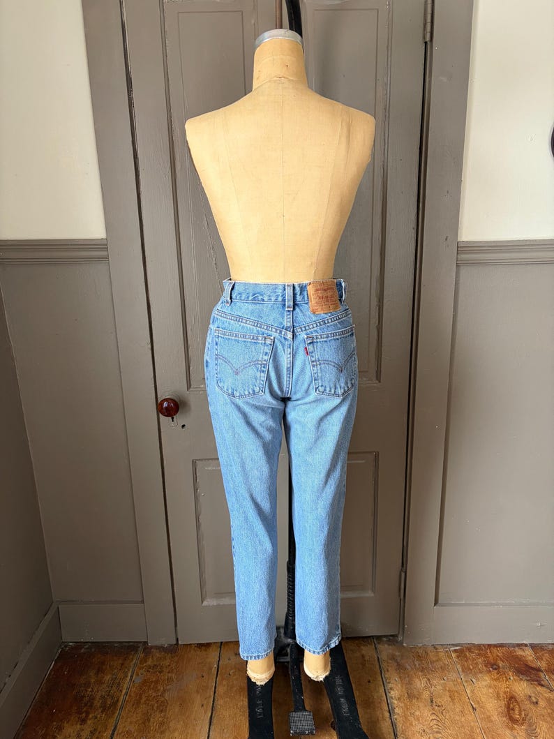 May include: Light blue denim jeans on a mannequin. The jeans have a classic five-pocket design, a button and zipper closure, and a leather patch on the back. The jeans are straight-legged and appear to be vintage.