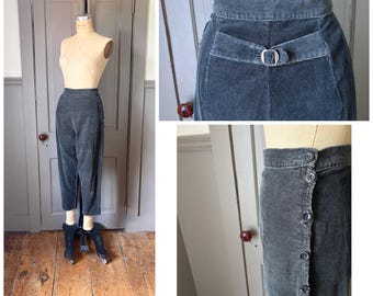 May include: Dark charcoal gray corduroy trousers featuring a high-waisted fit, a side button closure with five visible buttons, and a rear cinch strap with a silver-toned metal buckle. The pants have a tapered leg silhouette and a textured finish.