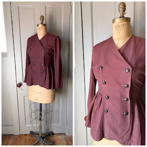 May include: A deep burgundy double-breasted jacket featuring long sleeves, a V-neckline, and a peplum waist. The garment is secured with eight dark buttons arranged in two vertical rows. The jacket is displayed on a beige dress form.