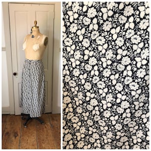 May include: A long, flowing skirt with a navy blue background and a white floral pattern. The skirt is displayed on a mannequin, showcasing its full length and design. The fabric's intricate floral print is also shown in a close-up view.