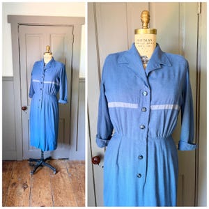 May include: A blue midi-length dress featuring a collared neckline, long sleeves with cuffs, and a horizontal light gray stripe across the chest. The dress has a button-down front closure with dark buttons and a gathered waistline on a mannequin.