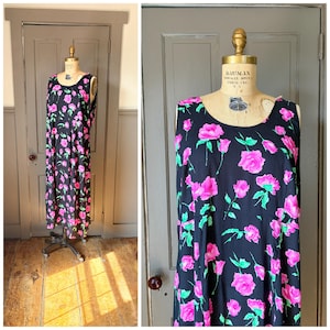 May include: A sleeveless floor-length dress in black featuring a repeating floral pattern of vibrant pink roses with green leaves. The garment is displayed on a beige dress form with a round neckline and a loose, flowing silhouette.