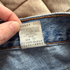May include: Close-up of a blue denim garment with a white tag. The tag has black text with washing instructions and numbers. The garment has orange stitching.