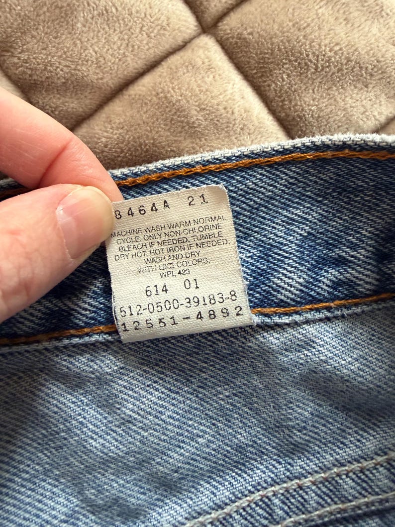 May include: Close-up of a blue denim garment with a white tag. The tag has black text with washing instructions and numbers. The garment has orange stitching.