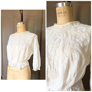 May include: Cream-colored blouse with intricate white floral embroidery. The long-sleeved top features a high, lace-trimmed collar and a ruffled hem. The blouse is displayed on a vintage dress form.