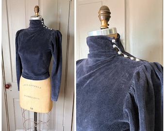 May include: A dark navy blue velvet long-sleeve top featuring a high mock neck collar. The garment includes a row of small white decorative buttons extending from the collar down the shoulder seam, with puffed sleeves and a fitted waist.