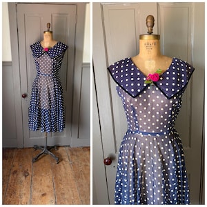 May include: Navy blue polka dot dress with a large collar and a pink flower accent. The dress is displayed on a vintage dress form. The dress features a sheer overlay and a flared skirt, creating a classic vintage look.