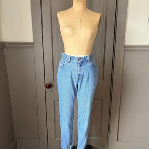 May include: Light blue denim jeans on a vintage dress form. The jeans have a classic five-pocket design and a button and zipper closure. The jeans are cuffed at the ankles. The dress form is made of a light-colored material and is positioned in front of a wooden door.