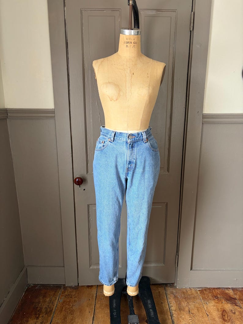 May include: Light blue denim jeans on a vintage dress form. The jeans have a classic five-pocket design and a button and zipper closure. The jeans are cuffed at the ankles. The dress form is made of a light-colored material and is positioned in front of a wooden door.