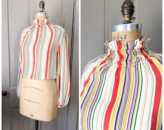 May include: A vintage blouse with vertical stripes in red, yellow, green, blue, and purple on a white background. The blouse has long sleeves and a high, ruffled neckline. The garment is displayed on a dress form.