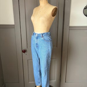 May include: Light blue denim jeans displayed on a vintage dress form. The jeans have a classic five-pocket design, a button and zipper closure, and a straight leg fit. The jeans are a medium wash and appear to be in good condition.
